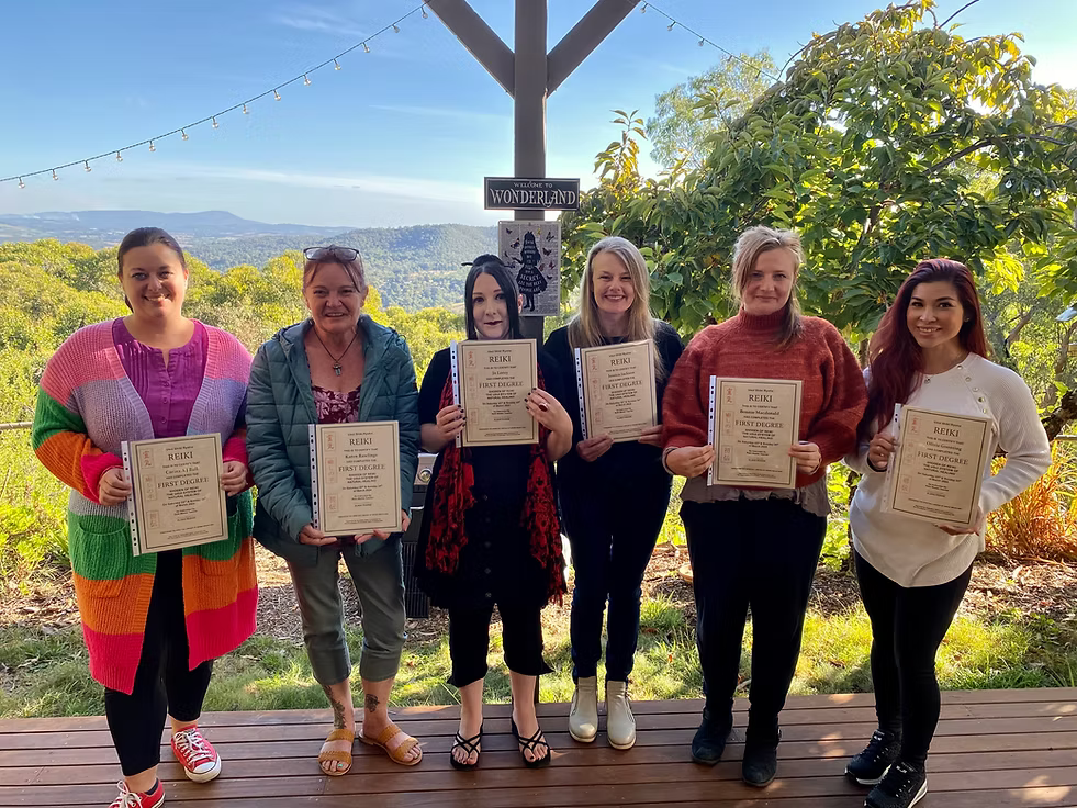 Six Reiki Level 1 graduates proudly holding their certificates on the deck at Earth Aya Energy, Healesville, with panoramic Yarra Valley views and blue skies in the background