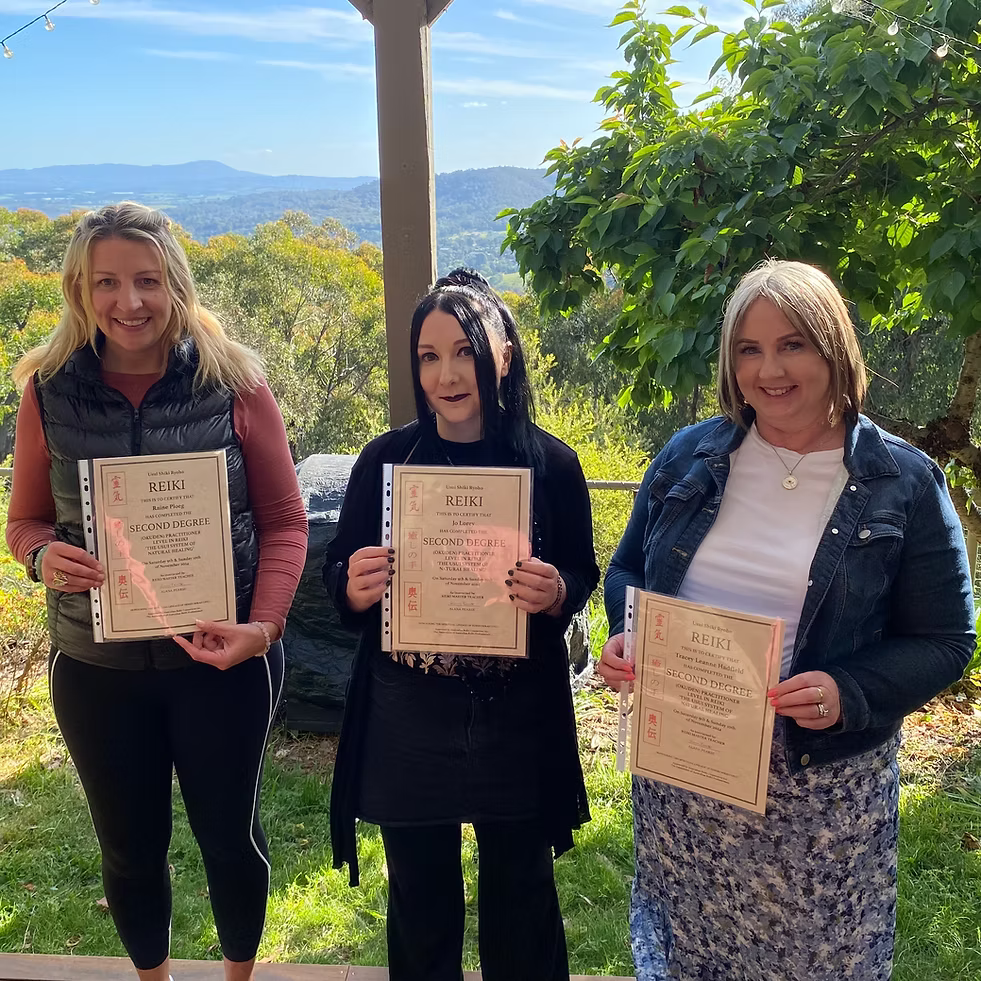 Three Reiki Level 2 graduates proudly holding their certificates in the garden at Earth Aya Energy, Healesville, with Yarra Valley views and blue skies in the background